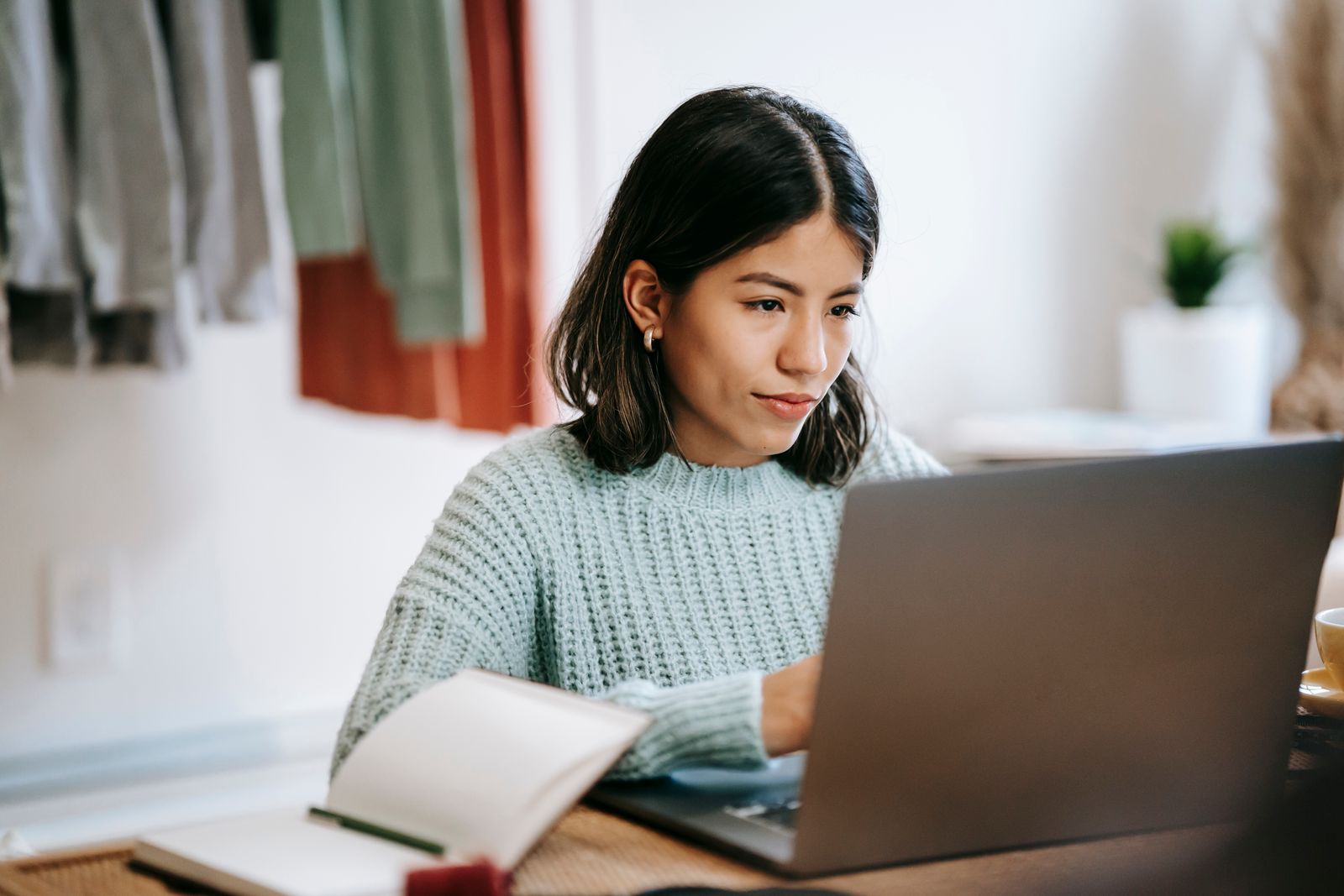 Una mujer haciendo un curso en una computadora port&aacute;til en la mesa de su casa, con un cuaderno abierto al lado.