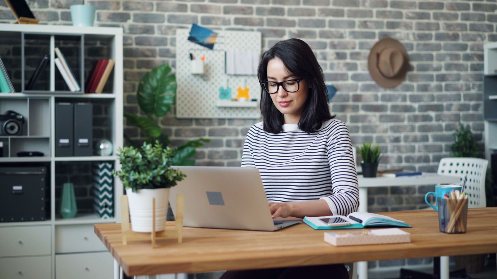 A young woman typing on a laptop at a wooden desk at home, with an open notebook beside her.